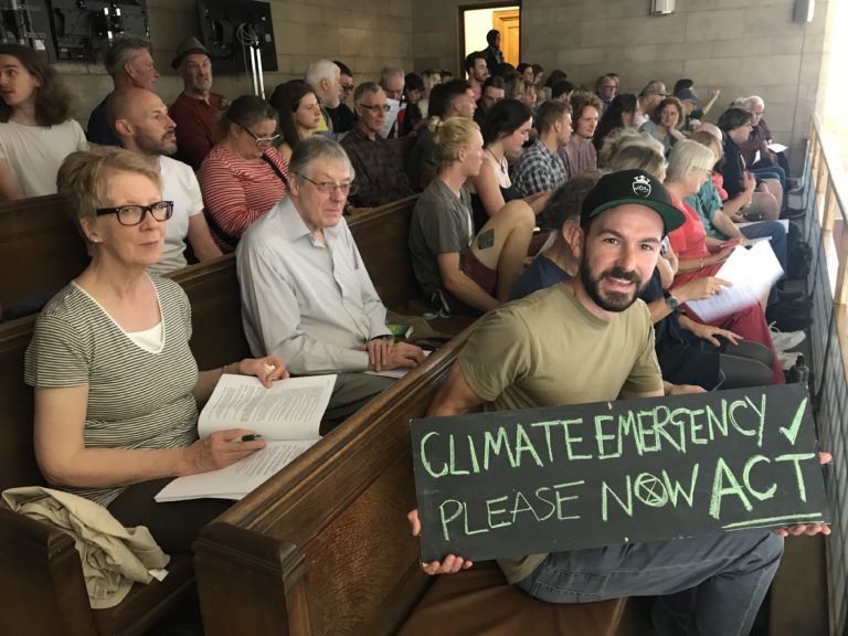 Campaigners at town meeting, one holding a sign saying Climate Emergency Please Now Act